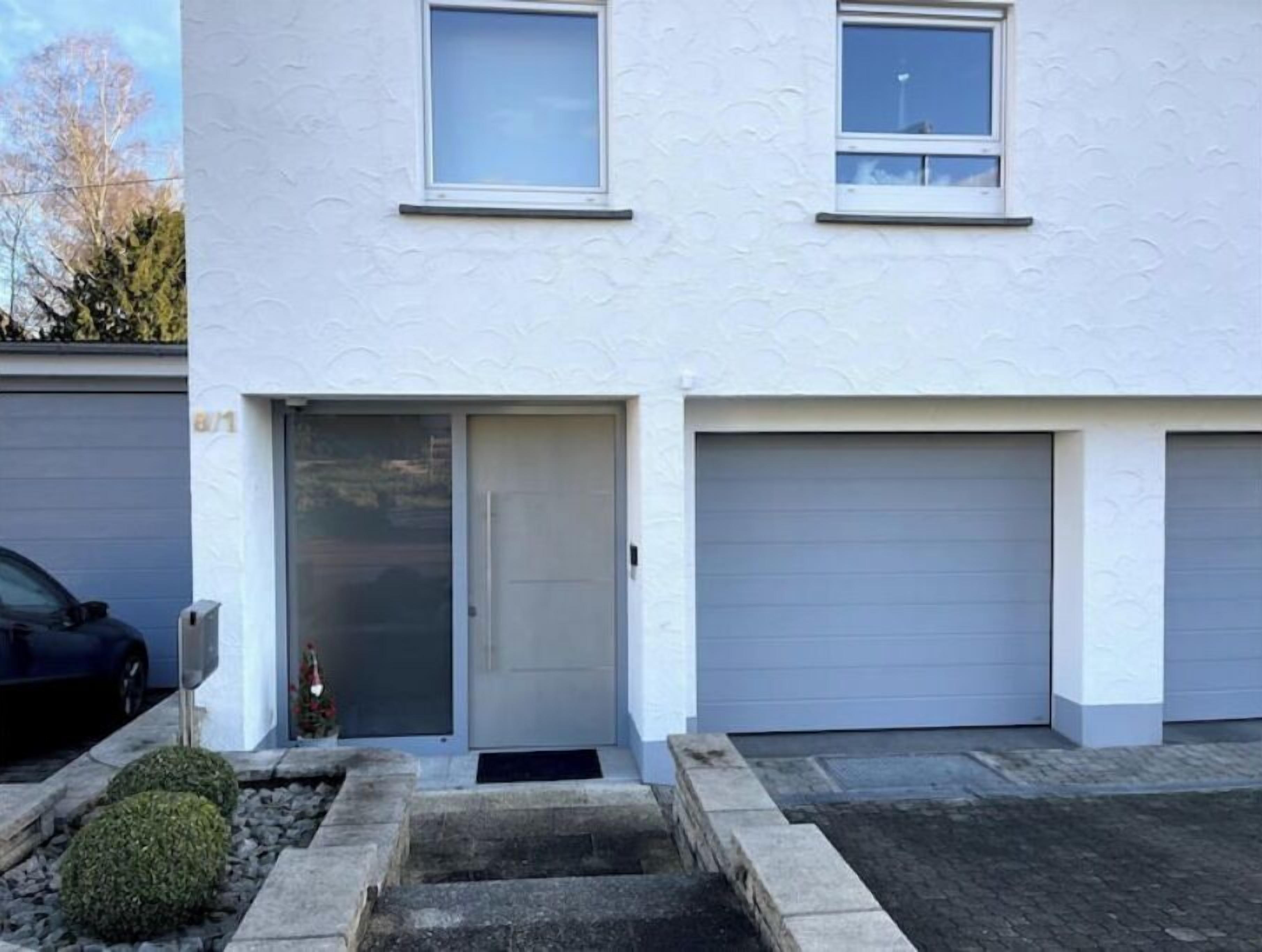 Een modern gebouw met twee verdiepingen, een garagepoort, een kleine ingang met een deur en een auto ervoor geparkeerd.