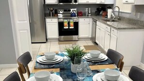 Cozinha moderna com armários brancos, eletrodomésticos de aço inoxidável e um espaço de refeições para seis pessoas, com toalha de mesa azul.