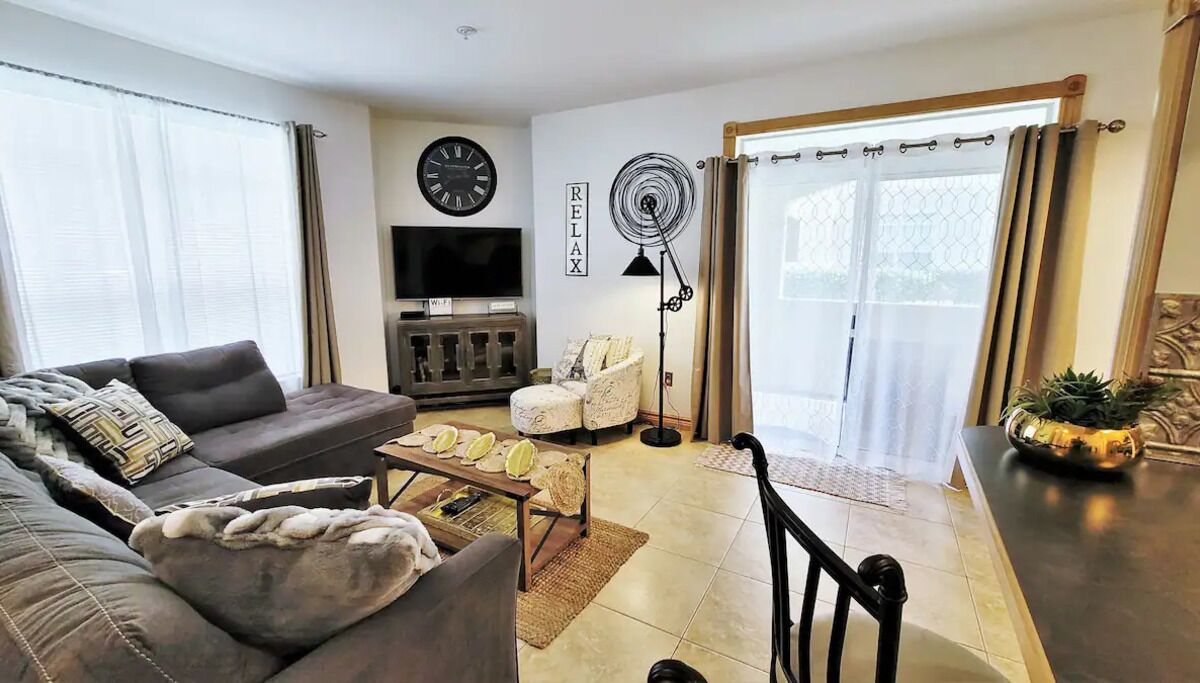 Una sala de estar con sofá, sillón, mesa de centro y televisor. Hay una chimenea, un ventilador y una ventana con cortinas.