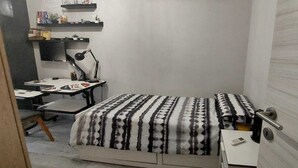 A small, neatly arranged bedroom with a bed, a desk, and a chair. There are shelves above the desk holding various items. - Roman Holidays (Roma)