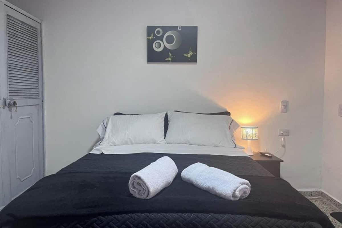 A neatly made bed with two rolled towels, a wall-mounted abstract art piece, and a bedside lamp.