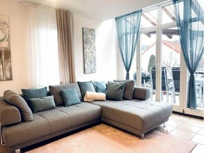Un salon moderne avec un canapé d’angle gris, un tapis beige et une grande fenêtre agrémentée de rideaux bleus.