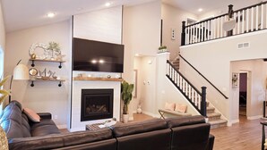 A spacious living room with a fireplace, a black leather sofa, a TV mounted on the wall, and a staircase leading to an upper level. - Posh Pelican House is guest favorite in quaint resort town of Breezy Point! (Breezy Point)
