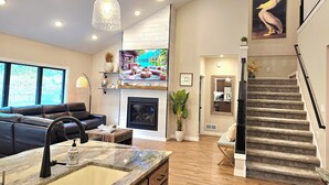 A spacious living area with a fireplace, a modern kitchen with a marble countertop, a staircase, and a large window offering a view of the outdoors. - Posh Pelican House is guest favorite in quaint resort town of Breezy Point! (Breezy Point)