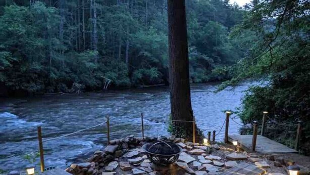A stone fire pit surrounded by wooden benches, set against a backdrop of a forested river. - Fun on the River - Swim, Fish, Hike Near Tubing (Blue Ridge)
