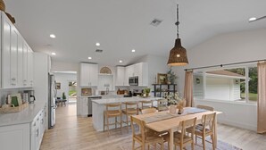 A modern kitchen with white cabinetry, a central island, and a dining area with wooden chairs. - Luxe Golden Hour - Private Pool & Pickleball Court, Outdoor Kitchen (Palm Beach Gardens)