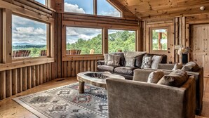 Een gezellig cabin interieur met houten details, grote ramen met een prachtig uitzicht, een leren bank en een vloerkleed met een patroon.