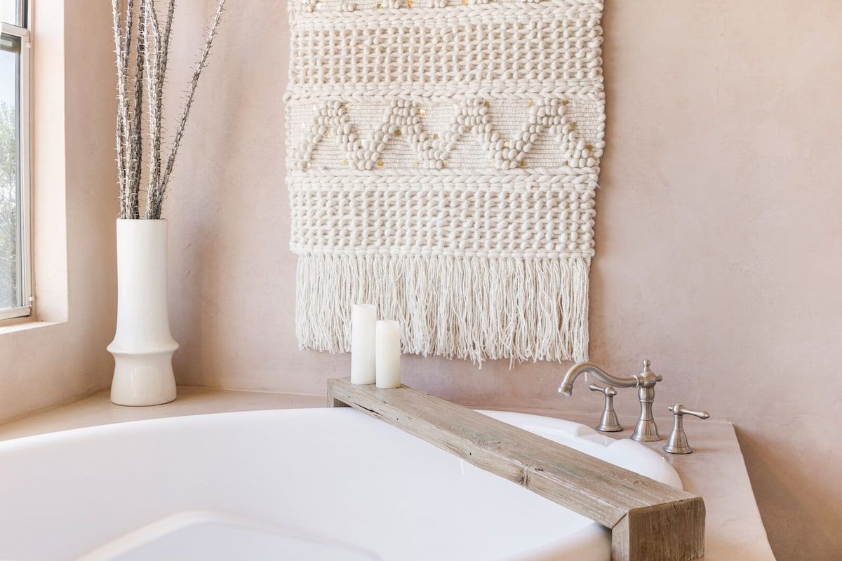 A bathroom with a white bathtub, a wooden sideboard, a macramé wall hanging, and a vase with branches.