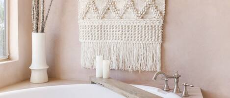 A bathroom with a white bathtub, a wooden sideboard, a macramé wall hanging, and a vase with branches.
