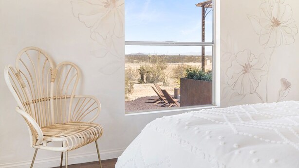 A room with a bed, a wicker chair, and a window with a view of a desert landscape. - Desert Moon-Hot Tub & Views Near Joshua Tree! (Joshua Tree)