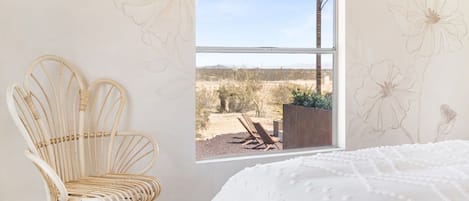 Una habitación con una cama, un sillón de mimbre y un ventanal con vistas a un paisaje desértico.