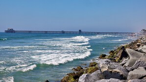 A coastal scene with a pier extending into the ocean, rocky shoreline, and waves crashing onto the beach. - Steps to the Sand | Modern Oceanfront Condo, A/C, Dog-Friendly, Walk to Pier (Oceanside)