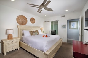 Quarto com cama, mesa de cabeceira, abajur, ventilador de teto e obras de arte nas paredes.