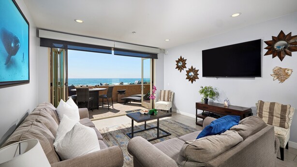 A modern living room with a large sofa, a coffee table, and a TV mounted on the wall. There is a balcony with a view of the ocean. - Steps to the Sand | Modern Oceanfront Condo, A/C, Dog-Friendly, Walk to Pier (Oceanside)