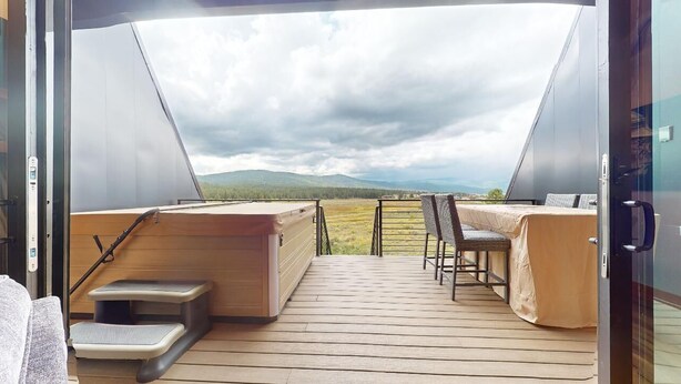A modern, open-plan room with a wooden deck, a hot tub, a covered seating area, and a view of the countryside.