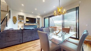 A modern living room with a glass dining table, grey sofa, and a large window offering a view of the outdoors.