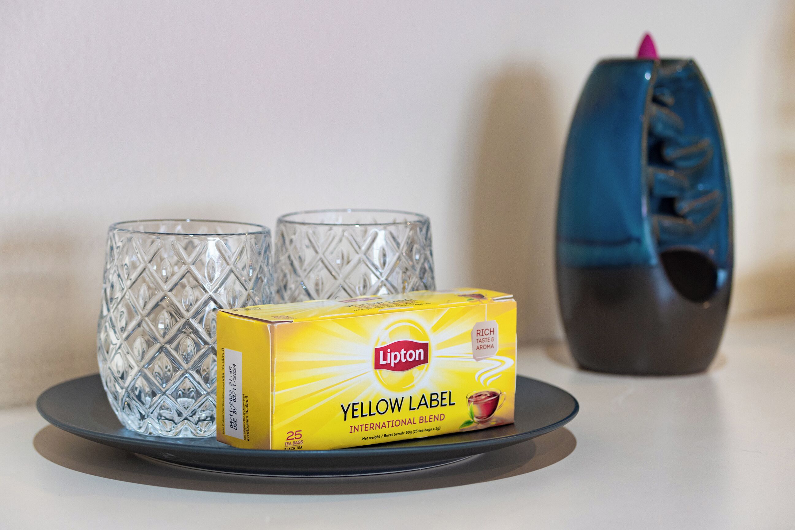 A box of Lipton Yellow Label International Blend tea, two clear glass cups, and a dark-colored vase on a white surface.