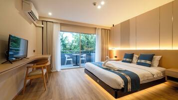 A modern hotel room with a large bed, a desk, a television, and a balcony with a view of greenery.