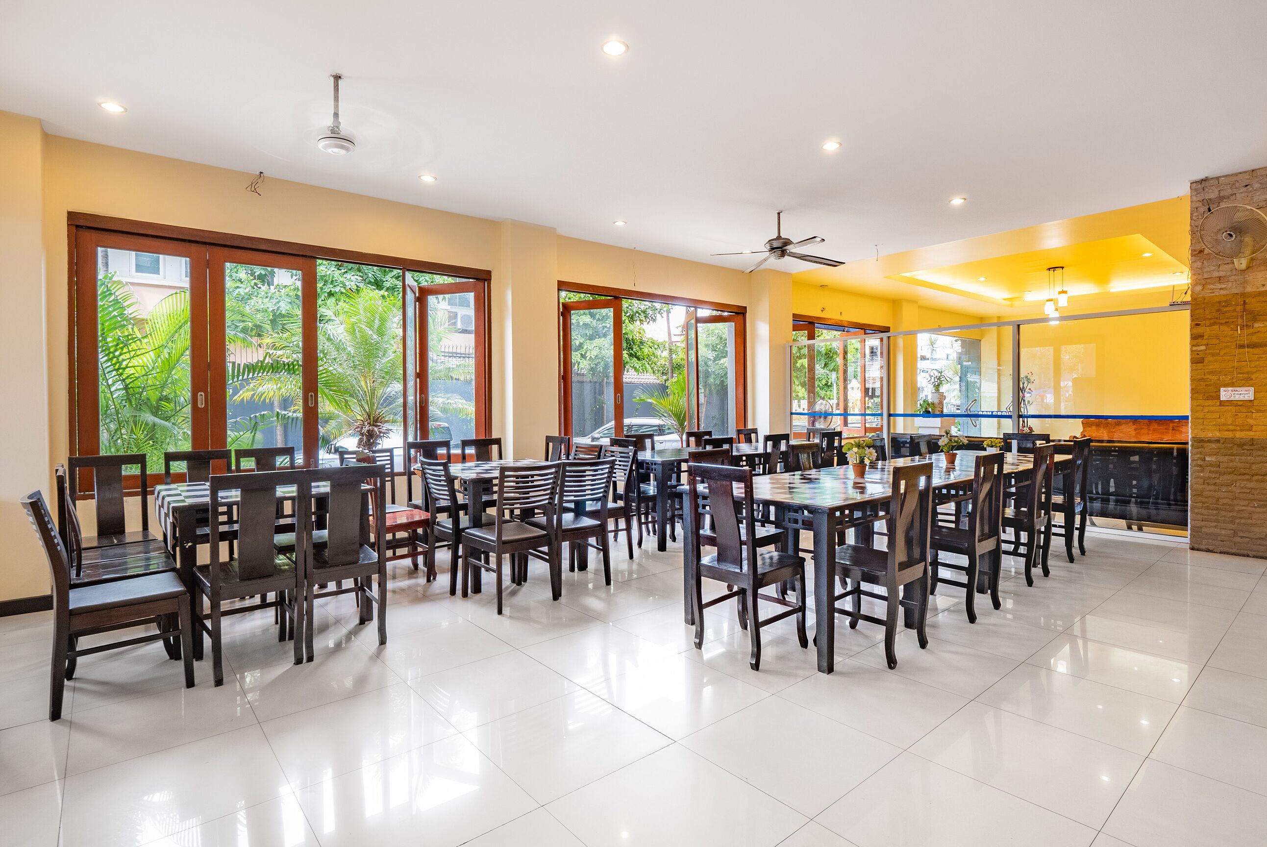 A spacious dining area with multiple tables and chairs, large windows with a view of greenery, and ceiling fans.