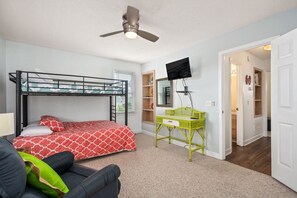 A bedroom with a bunk bed, a desk with a chair, a ceiling fan, a TV mounted on the wall, and a door leading to another room. - Sandy T'z: Soundside, Golf Course Views, Oceanfront Community Pool (Nags Head)