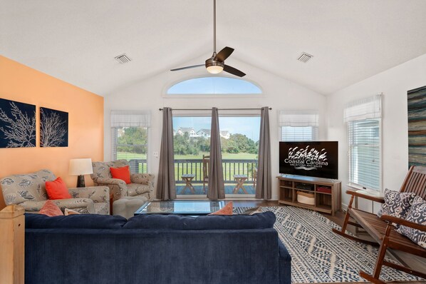 A spacious living room with a large sofa, a coffee table, and a television. There is a balcony with a view of the greenery and houses. - Sandy T'z: Soundside, Golf Course Views, Oceanfront Community Pool (Nags Head)