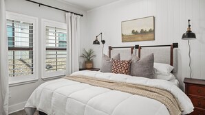 A bedroom with a large bed, a headboard with leather straps, a bedside table with a lamp, and a window with blinds.