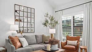 A living room with a grey sofa, a wooden coffee table, a brown leather armchair, and a small wooden stool. There is a mirror on the wall, a potted plant, and a window with curtains.