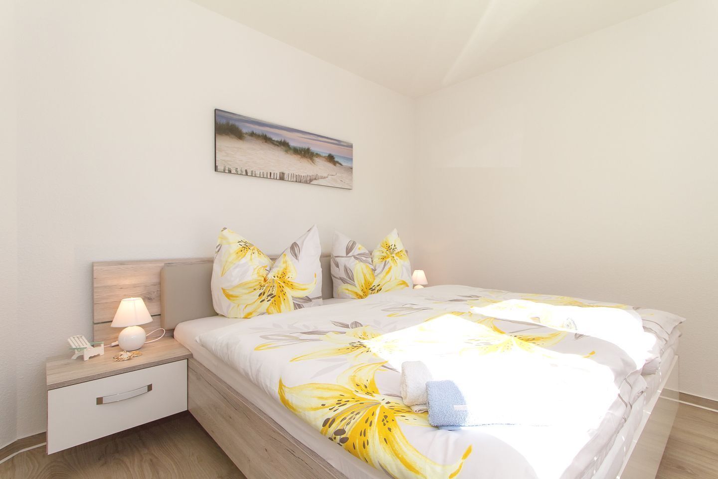 Un dormitorio con cama, mesitas de noche, lámpara, un cuadro y suelo de madera.