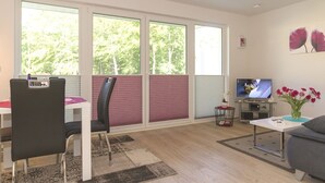 A modern living room with a dining area, large windows, a flat-screen TV, and a vase of flowers.