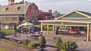 A covered outdoor seating area with tables and chairs, a red building with a green roof in the background, and a stone wall in the foreground.