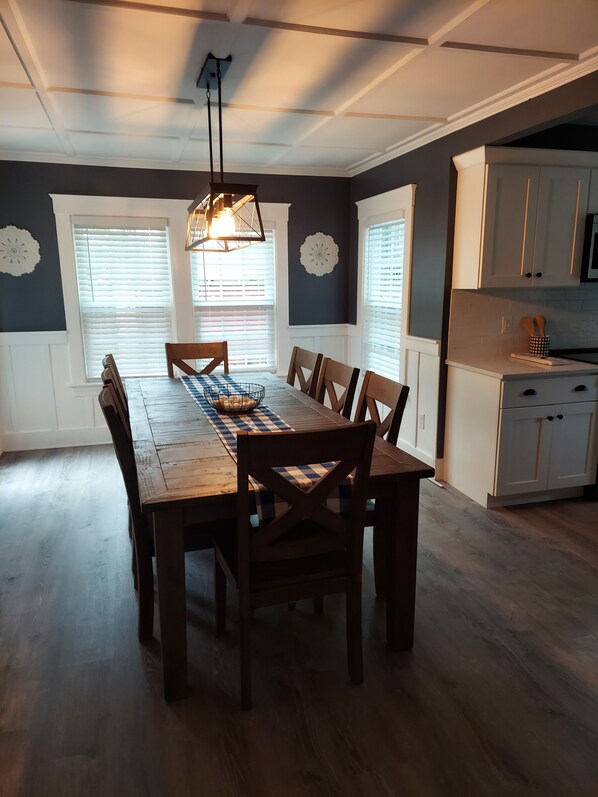 A dining area with a wooden table and chairs, a hanging light fixture, and a kitchen with white cabinets.