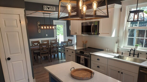 A modern kitchen with a central island, a dining area with wooden chairs, and a view of the outdoors through a window.