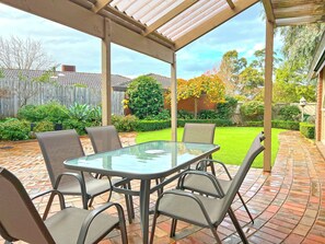 Ein überdachter Patio-Bereich mit Glastisch und Metallstühlen, umgeben von einer Ziegelterrasse und einem gepflegten Rasen.