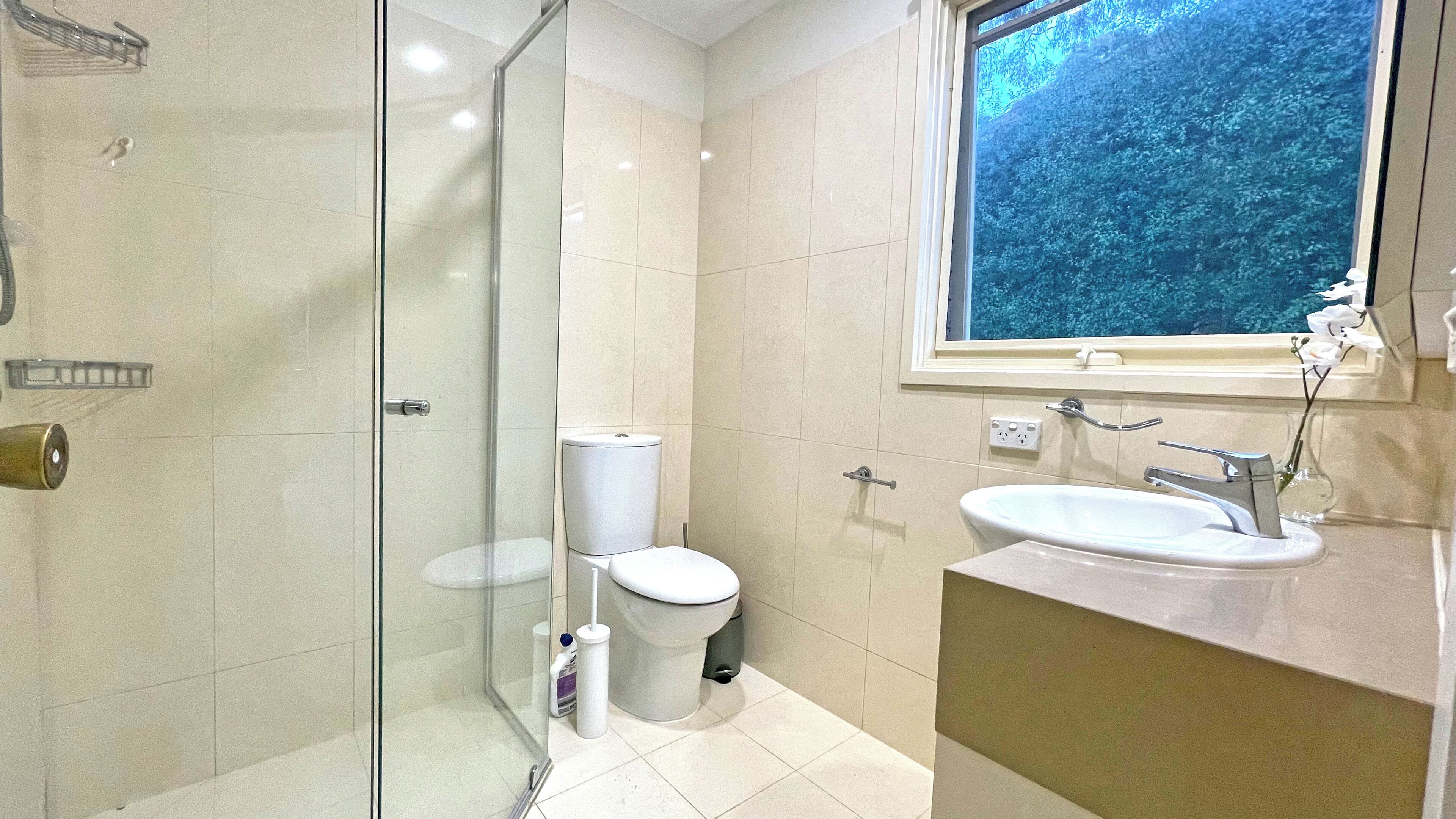 A modern bathroom with a glass-enclosed shower, a toilet, and a sink with a window above.