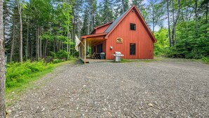 Une cabane en bois rouge, dotée d’un porche, entourée d’une allée de gravier et d’une forêt dense.