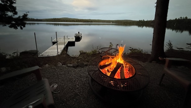 Un feu de camp au bord du lac, avec une jetée qui s’avance dans l’eau, entouré d’arbres et sous un ciel dégagé.