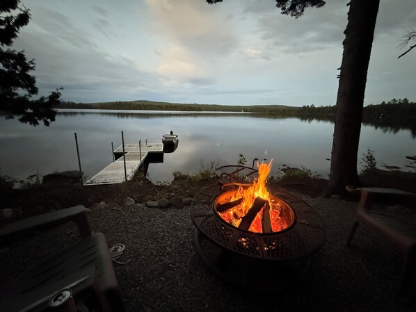 A lakeside campfire with a dock extending into the water, surrounded by trees and a clear sky.