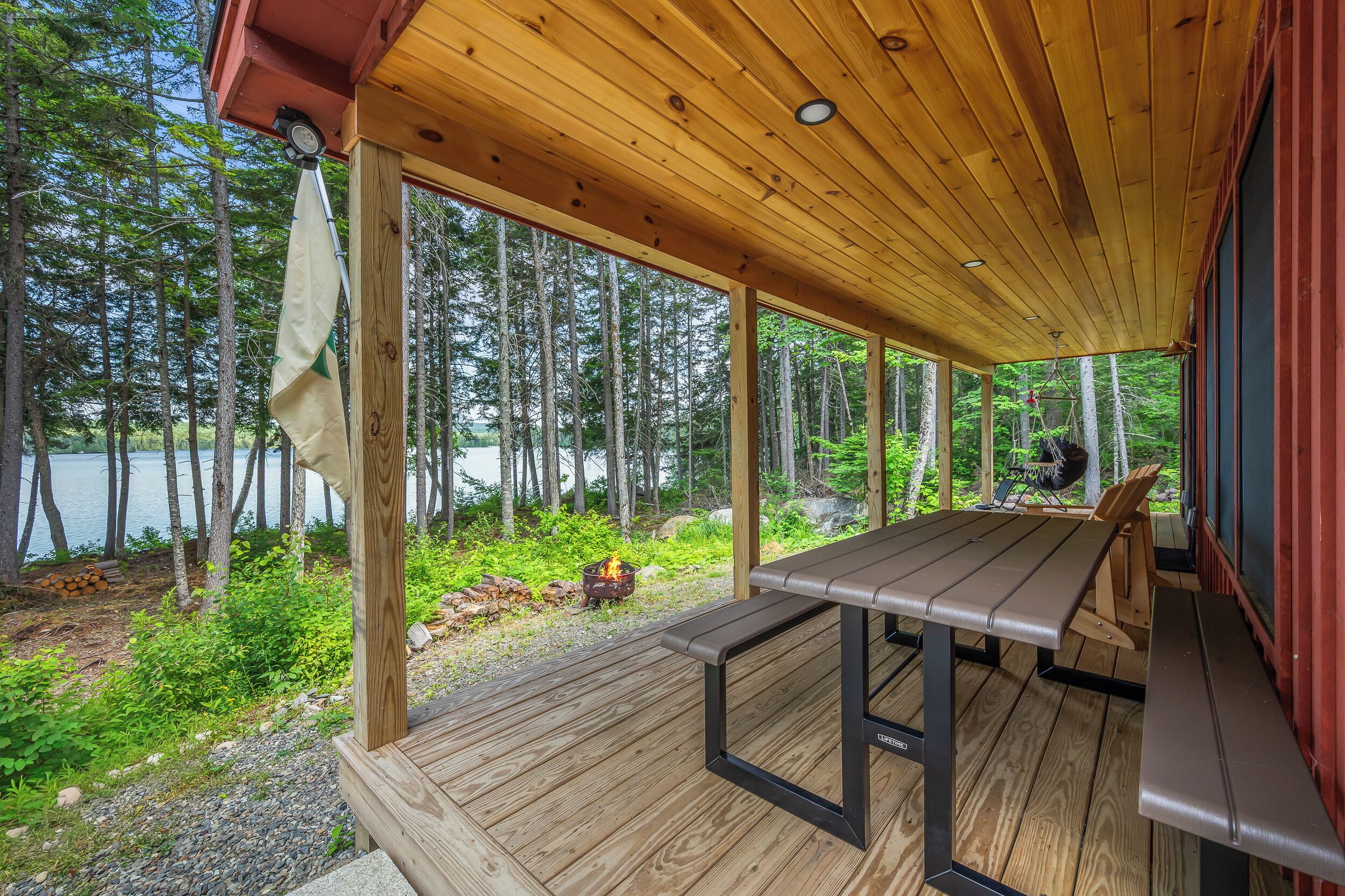 A covered outdoor seating area with a table and benches, overlooking a forested area and a body of water.