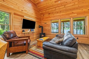 A cozy cabin interior with a leather sofa, a wooden chair, a flat-screen TV mounted on the wall, and a fireplace. - Richardson Pond - rare opportunity to rent on this body of water! (Rangeley)