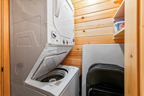 A compact, modern laundry room with a built-in washer and dryer, wooden panel walls, and a small seating area with a chair.