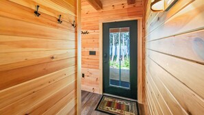 Un couloir intérieur en bois, avec une porte donnant sur l’extérieur, un luminaire fixé au mur et un paillasson.