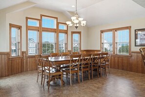 Comedor con una gran mesa de madera y sillas, ventanas amplias con persianas y una lámpara de araña.