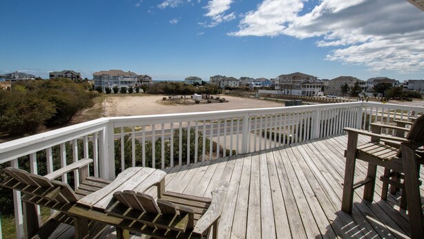Een houten terras met een witte reling met uitzicht op een strand en huizen.