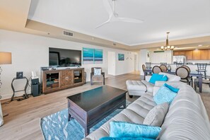 A spacious living room with a large sofa, a coffee table, and a TV. There is a dining area with a table and chairs, and a kitchen in the background. - Welcome to Dolphin Kisses and Sea Turtle Hearts! Harbor Landing 101B! (Destin)