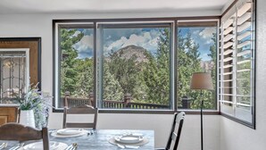 Zona pranzo con tavolo apparecchiato per due, una finestra vista alberi e montagna, e una lampada da terra.