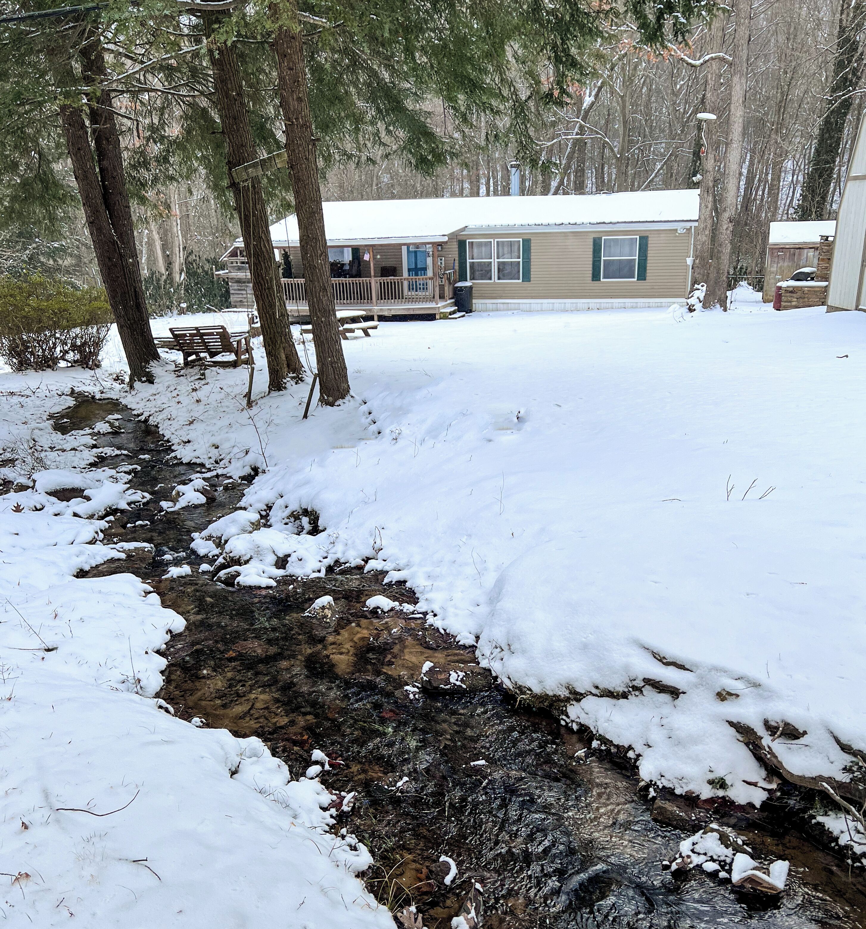 Un espace extérieur enneigé avec un banc en bois, un petit ruisseau et une maison en arrière-plan.