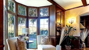 A room with a large bay window, wooden flooring, two upholstered chairs, a small table with a lamp, and a vase with plants. - 5-Min Walk To Rose Bowl - Luxe Estate! (Pasadena)