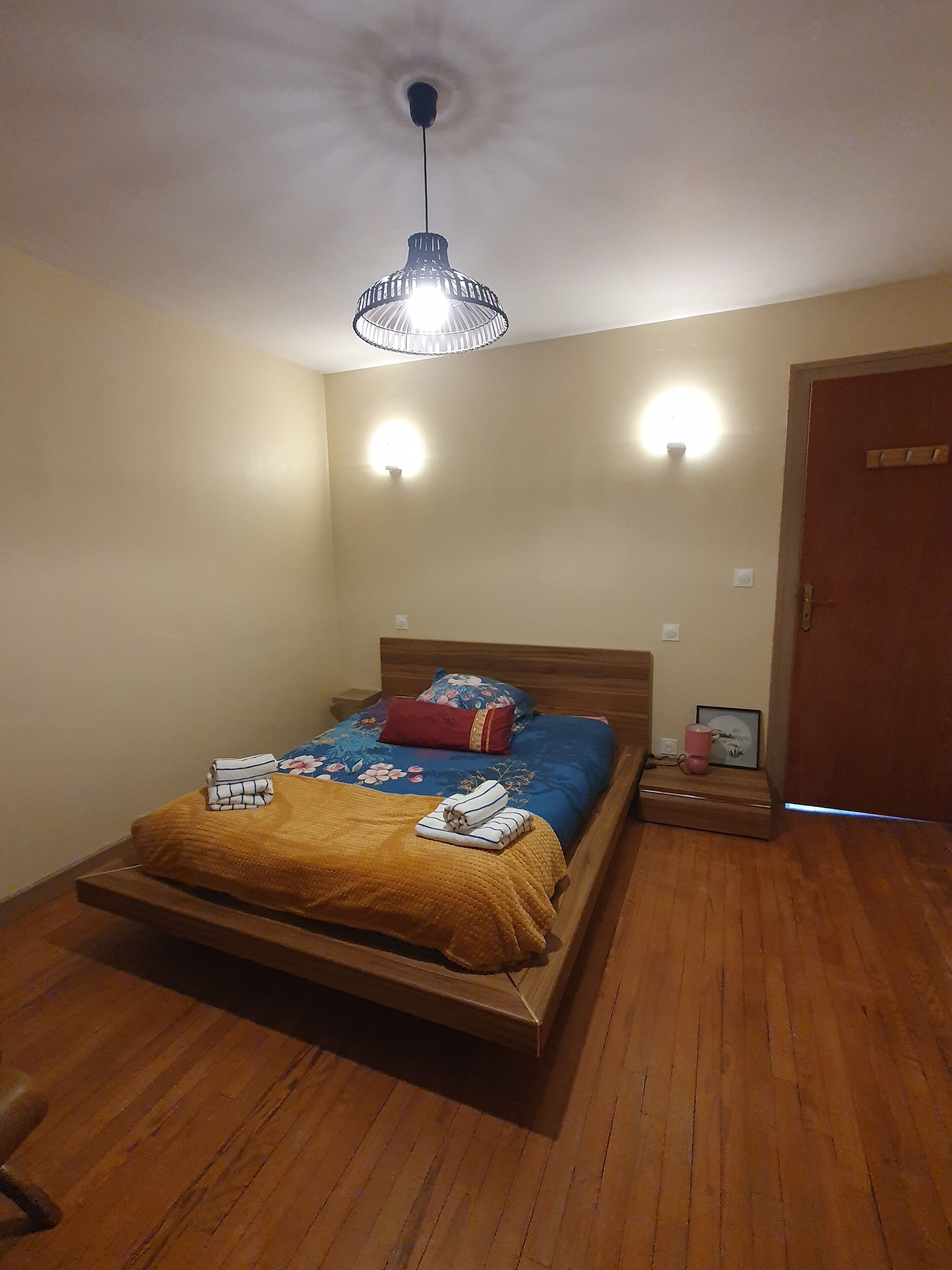 A single bed with a wooden frame, a blue and red floral bedspread, and a yellow throw blanket. There are two wall-mounted lights and a ceiling light fixture.