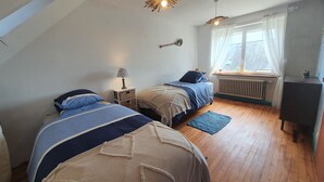 Ein Schlafzimmer mit zwei Betten, einem Fenster mit Vorhängen, einem Heizkörper und einem kleinen Tisch mit einer Lampe.