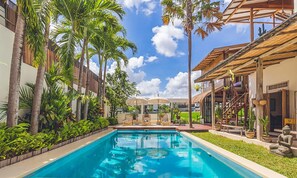 A swimming pool surrounded by palm trees and lounge chairs, with a covered outdoor seating area and a building with a thatched roof.